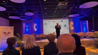 Mężczyzna w garniturze przemawia do mikrofonu na scenie, pełniąc rolę moderatora wydarzenia, co potwierdza napis na slajdzie „BARTŁOMIEJ PIECHACZEK”. Po bokach sceny ustawione są żółte fotele i banery informacyjne. Zdjęcie wykonane z perspektywy widza siedzącego w jednym z pierwszych rzędów.