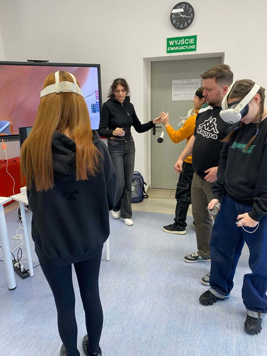 Grupa osób w jasnej sali lekcyjnej podczas warsztatów. Dwie dziewczyny mają na głowach białe gogle VR i trzymają kontrolery. Obok nich stoi dwóch instruktorów, którzy pomagają w obsłudze sprzętu. Nad drzwiami widoczny jest zegar oraz zielony znak „Wyjście ewakuacyjne”.