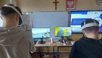 Dwóch uczniów stoi tyłem, prezentując gogle VR na głowach podczas pracy z systemem symulacji. Przed nimi znajdują się dwa monitory wyświetlające różne perspektywy wirtualnego środowiska pracy. W tle widać prezentację multimedialną dotyczącą Krajowego Planu Odbudowy i funduszy europejskich. Na biurku pod monitorami leżą dodatkowe akcesoria i pudełka ze sprzętem.