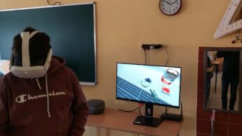 Zbliżenie na ucznia w brązowej bluzie z kapturem, który ma założone białe gogle VR. Obok na biurku stoi monitor wyświetlający realistyczną grafikę 3D z puszką farby i narzędziami. W tle widać zieloną tablicę szkolną oraz dużą ekierkę geometryczną zawieszoną na ścianie.