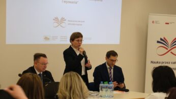 Zbliżenie na kobietę przemawiającą do mikrofonu, ubraną w ciemny żakiet i białą koszulę. Siedzący obok niej mężczyzna w okularach patrzy w dół, trzymając w dłoniach telefon. Na stole przed nimi znajdują się szklanki, butelki z wodą oraz czarny laptop.