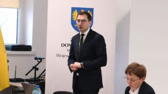 Mężczyzna w okularach i ciemnym garniturze stoi i przemawia, trzymając splecione dłonie przed sobą. Obok niego, przy stole, siedzi kobieta skupiona na notatkach lub dokumentach. W tle znajduje się biały baner z herbem oraz okno, przez które wpada naturalne światło.