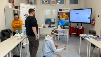 Wnętrze sali z uczestnikami warsztatów technologicznych. Chłopak w szarej bluzie klęczy na podłodze z goglami VR na oczach i kontrolerem w dłoni. W tle stoją dwaj mężczyźni obserwujący jego działania, a na stołach i ścianach widoczne są monitory, banery informacyjne „Europe Direct” oraz pomoce naukowe (model anatomiczny).