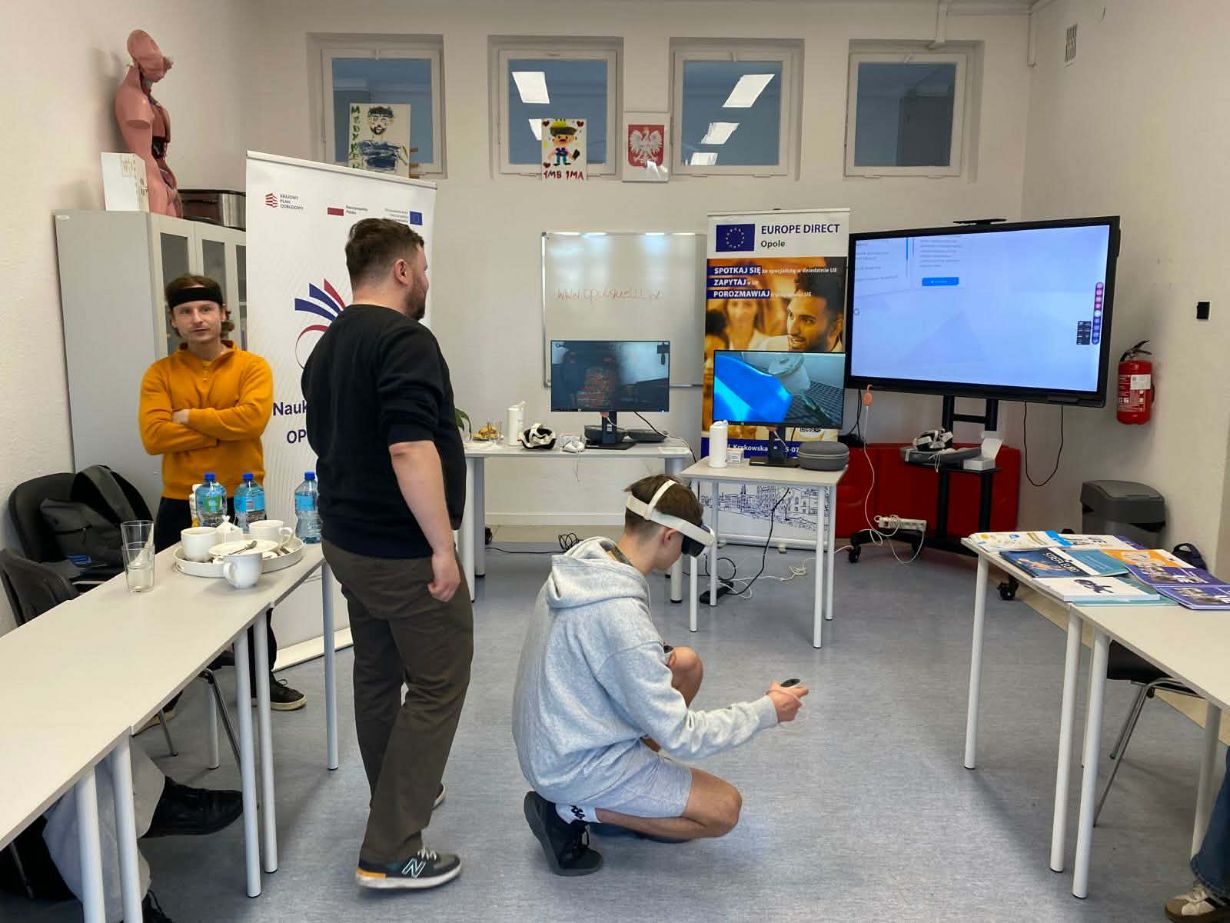 Wnętrze sali z uczestnikami warsztatów technologicznych. Chłopak w szarej bluzie klęczy na podłodze z goglami VR na oczach i kontrolerem w dłoni. W tle stoją dwaj mężczyźni obserwujący jego działania, a na stołach i ścianach widoczne są monitory, banery informacyjne „Europe Direct” oraz pomoce naukowe (model anatomiczny).