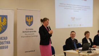 Kobieta w różowej sukience i czarnym żakiecie stoi przy banerze wicemarszałek województwa i przemawia do mikrofonu. Obok niej przy stole siedzą uśmiechnięty mężczyzna w garniturze oraz kobieta w okularach. W tle widoczny jest fragment ekranu z tytułem debaty.