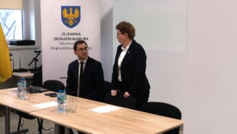 Kobieta w ciemnej marynarce stoi przy stole konferencyjnym, patrząc w stronę siedzącego obok mężczyzny w okularach. Oboje znajdują się w sali wykładowej przed jasnym ekranem projekcyjnym i banerem z napisem 