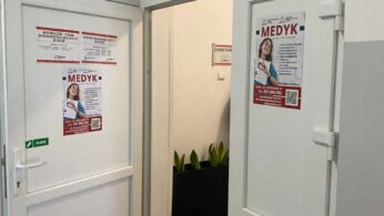Otwarte białe drzwi prowadzące do pomieszczenia z napisem „MEDYK” umieszczonym nad futryną. Na skrzydłach drzwi oraz obok nich naklejone są plakaty informacyjne szkoły policealnej oraz liceum ogólnokształcącego w Nysie. W głębi widać fragment korytarza z roślinami w donicach.