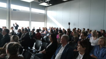 Podobne ujęcie siedzącej publiczności, ukazujące różnorodną grupę osób uczestniczących w prelekcji lub panelu dyskusyjnym. Na pierwszym planie widoczne są profile osób w ciemnych marynarkach i jasnych koszulach. Przez wysokie okna po lewej stronie wpada intensywne, dzienne światło.