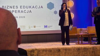Zbliżenie na kobietę przemawiającą do mikrofonu na scenie, ubraną w ciemną marynarkę i jasną koszulę. Obok niej stoi mężczyzna z podkładką na dokumenty, a w tle widać ekran z napisem „BIZNES EDUKACJA KOOPERACJA”. Scena jest oświetlona pionowymi, niebieskimi strumieniami światła.