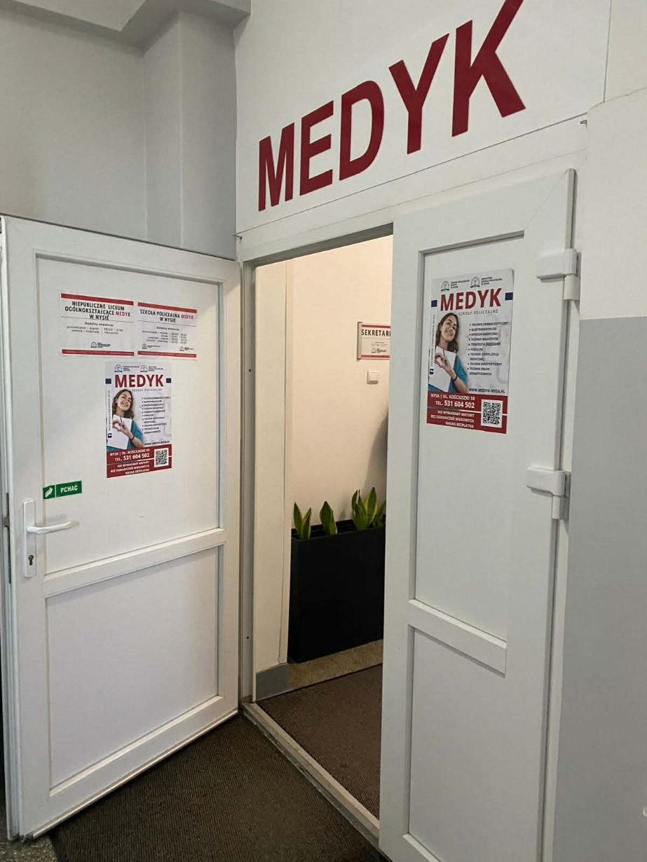 Otwarte białe drzwi prowadzące do pomieszczenia z napisem „MEDYK” umieszczonym nad futryną. Na skrzydłach drzwi oraz obok nich naklejone są plakaty informacyjne szkoły policealnej oraz liceum ogólnokształcącego w Nysie. W głębi widać fragment korytarza z roślinami w donicach.