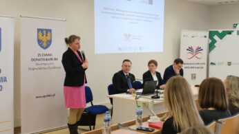 Szerokie ujęcie przedstawiające kobietę w różowej sukience przemawiającą do mikrofonu na tle oficjalnych banerów. Reszta panelistów siedzi przy stole, a jeden z mężczyzn podpiera głowę ręką. Na pierwszym planie widać rzędy ławek z uczestnikami oraz kubek kawy na jednym z blatów.