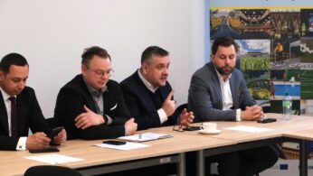 Czterej mężczyźni siedzą rzędem przy stole konferencyjnym, angażując się w rozmowę. Jeden z nich, w ciemnym swetrze, wykonuje gest wskazujący palcem, podkreślając swoją wypowiedź. Z prawej strony kadru widoczny jest fragment kolorowego baneru promocyjnego z napisem 