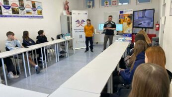 Grupa uczniów siedzi przy białych stołach ustawionych w podkowę, słuchając dwóch prowadzących stojących na środku sali. W tle znajdują się banery edukacyjne, duży monitor interaktywny oraz plansze na ścianach. Panuje atmosfera wykładu lub prezentacji.