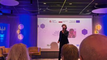 Młody mężczyzna z kręconymi włosami przemawia dynamicznie na scenie, a na ekranie za nim widnieje napis „FILIP HORNIK AKCJA UCZNIOWSKA”. Prelegent ma na sobie ciemny garnitur i gestykuluje jedną ręką. Na pierwszym planie widać nieostre zarysy głów uczestników wydarzenia.