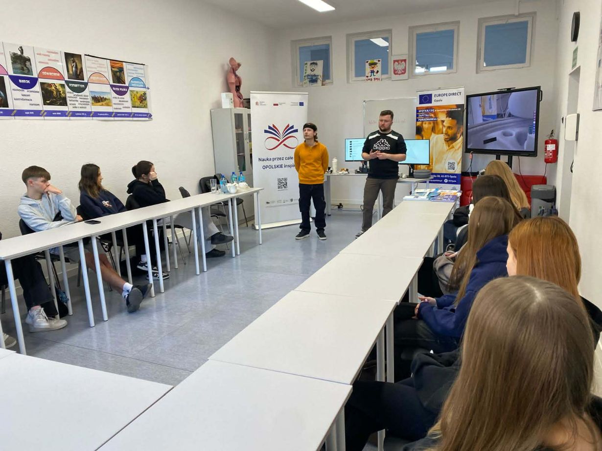 Grupa uczniów siedzi przy białych stołach ustawionych w podkowę, słuchając dwóch prowadzących stojących na środku sali. W tle znajdują się banery edukacyjne, duży monitor interaktywny oraz plansze na ścianach. Panuje atmosfera wykładu lub prezentacji.