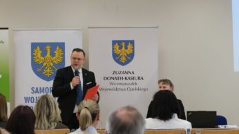 Mężczyzna w okularach i ciemnym garniturze stoi na podium, trzymając w rękach czerwony folder i przemawiając do mikrofonu. W tle widoczne są banery z herbem województwa opolskiego oraz nazwiskiem wicemarszałek. Na dole zdjęcia widoczne są tyły głów słuchaczy siedzących w sali.