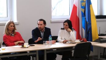 Trzy osoby siedzą przy stole konferencyjnym na tle okna i flag państwowych. Mężczyzna w środku gestykuluje prawą ręką podczas wypowiedzi, a siedzące obok kobiety słuchają go z uwagą. Na stole widać poczęstunek, napoje oraz przybory do pisania.