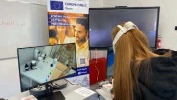 Zbliżenie na stanowisko z technologią VR. Widoczna od tyłu dziewczyna z długimi, rudymi włosami ma na głowie białe gogle. Przed nią stoi monitor, na którym wyświetlany jest podgląd z wirtualnej rzeczywistości (widok gabinetu medycznego/kosmetycznego). Obok znajduje się baner „Europe Direct Opole”.