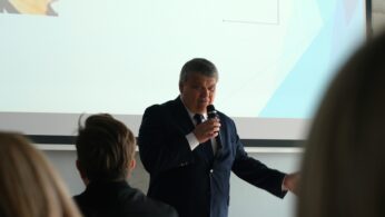 Portret przemawiającego mężczyzny w ciemnym garniturze i krawacie, trzymającego mikrofon przy ustach. W tle widoczny jest fragment slajdu z nagłówkiem „Opole, 11.03.2026”. Zdjęcie wykonane z perspektywy widza siedzącego w ostatnich rzędach, co sugerują rozmyte sylwetki przed mikrofonem.
