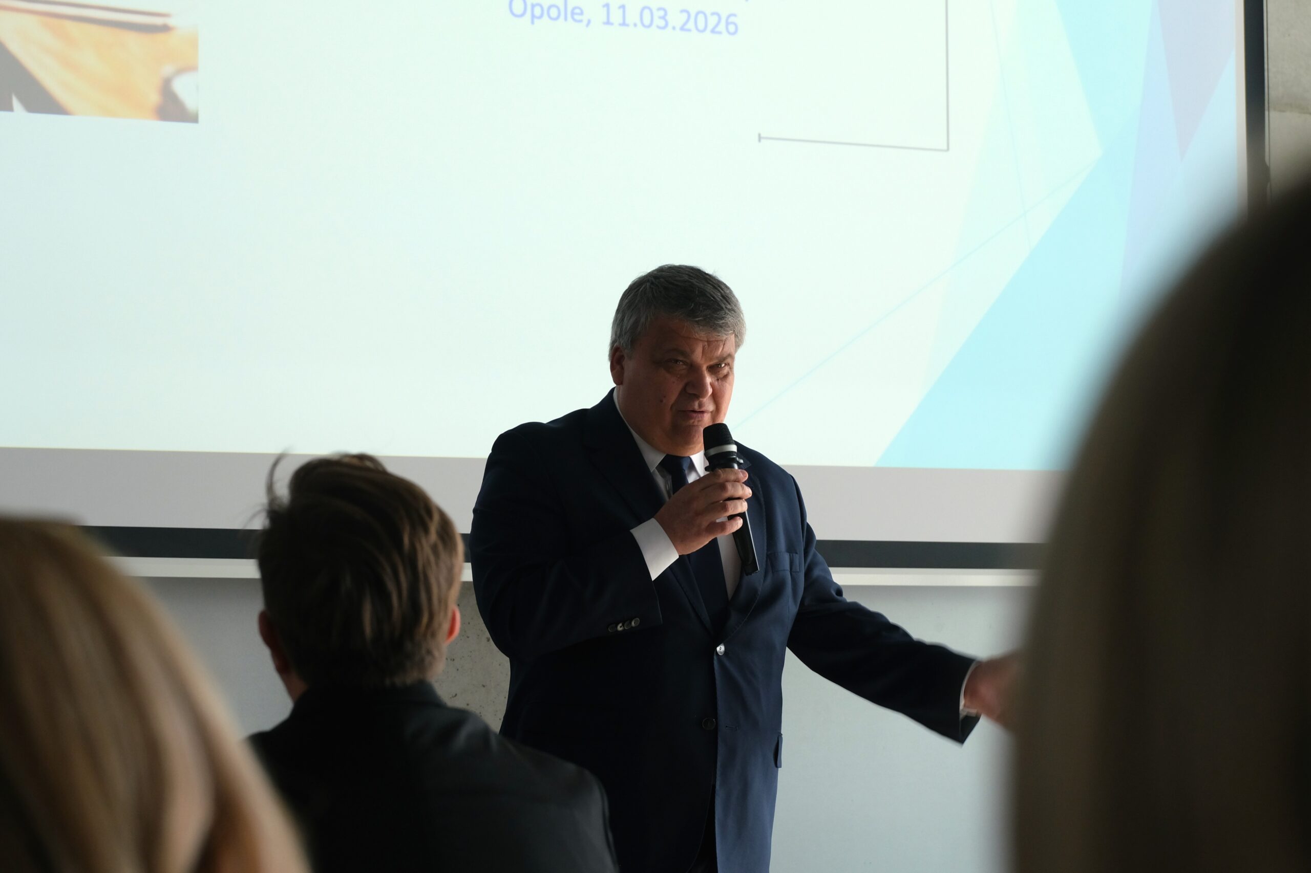 Portret przemawiającego mężczyzny w ciemnym garniturze i krawacie, trzymającego mikrofon przy ustach. W tle widoczny jest fragment slajdu z nagłówkiem „Opole, 11.03.2026”. Zdjęcie wykonane z perspektywy widza siedzącego w ostatnich rzędach, co sugerują rozmyte sylwetki przed mikrofonem.