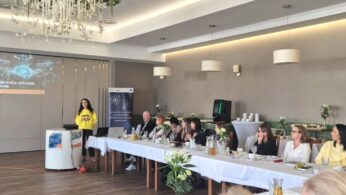 Prelegentka w jaskrawożółtej bluzie z napisem „matcha” stoi przy rzutniku, prowadząc wykład pt. „Psychologia w erze sztucznej inteligencji (AI)”. Przed nią siedzą uczestnicy spotkania, a na stole widać kwiaty w wazonach oraz baner projektu edukacyjnego.