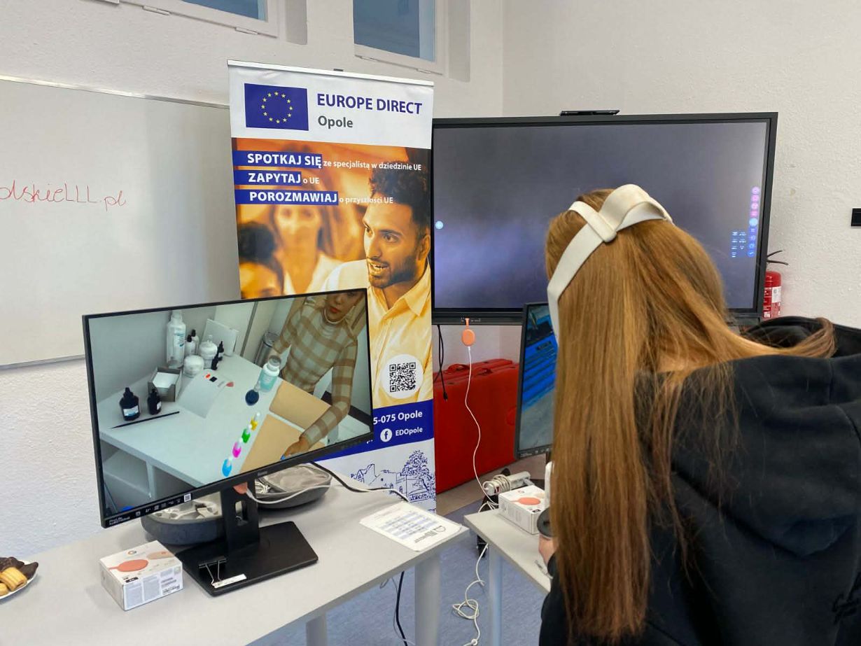 Zbliżenie na stanowisko z technologią VR. Widoczna od tyłu dziewczyna z długimi, rudymi włosami ma na głowie białe gogle. Przed nią stoi monitor, na którym wyświetlany jest podgląd z wirtualnej rzeczywistości (widok gabinetu medycznego/kosmetycznego). Obok znajduje się baner „Europe Direct Opole”.