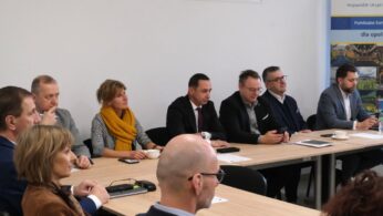 Szerokie ujęcie uczestników spotkania siedzących wzdłuż długiego stołu w jasnej sali. Na pierwszym planie widać tył głowy jednej z osób, a dalej rządy mężczyzn i kobiet obserwujących przebieg konferencji. W tle znajduje się duży baner z logotypami Funduszy Europejskich.