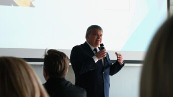 Mężczyzna w ciemnym garniturze kontynuuje swoje wystąpienie, gestykulując lewą ręką dla podkreślenia wypowiedzi. Światło z ekranu za jego plecami lekko prześwietla tło, skupiając uwagę na mówcy. Atmosfera sugeruje oficjalne wystąpienie eksperckie lub polityczne.