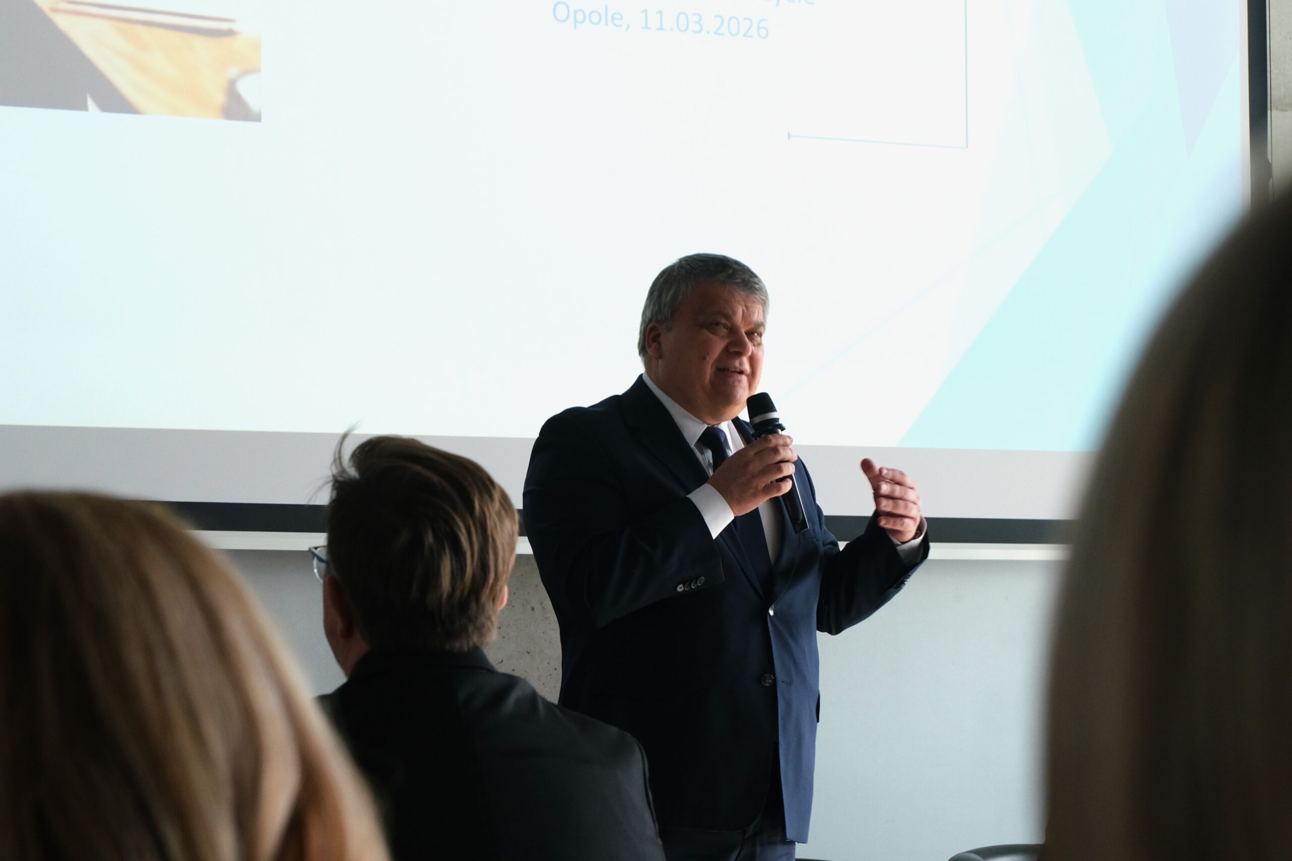 Mężczyzna w ciemnym garniturze kontynuuje swoje wystąpienie, gestykulując lewą ręką dla podkreślenia wypowiedzi. Światło z ekranu za jego plecami lekko prześwietla tło, skupiając uwagę na mówcy. Atmosfera sugeruje oficjalne wystąpienie eksperckie lub polityczne.