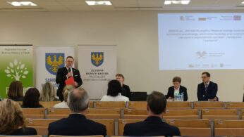 Widok z głębi sali na panelistów siedzących i stojących przy stole prezydialnym pod dużym ekranem. Jeden z mężczyzn przemawia, trzymając czerwony dokument, podczas gdy reszta grupy słucha. Sala jest jasna, z kasetonowym sufitem i rzędami drewnianych ławek dla publiczności.
