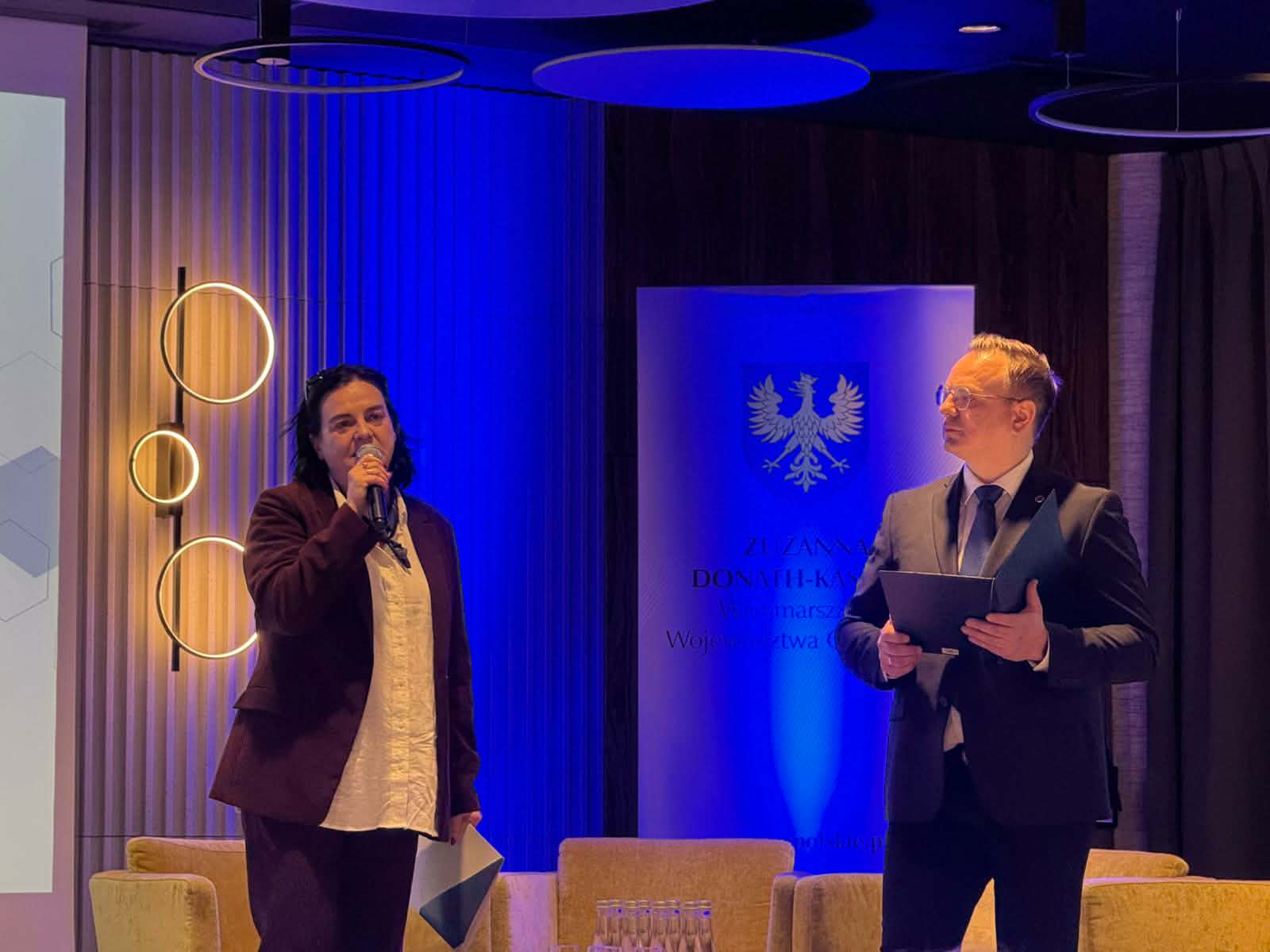 Portretowe ujęcie dwójki prowadzących na scenie podświetlonej na niebiesko. Kobieta mówi do mikrofonu, trzymając w dłoni ulotkę, podczas gdy mężczyzna obok niej spogląda w jej stronę, trzymając otwartą teczkę. W tle widoczny jest herb na niebieskim banerze.