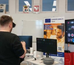 Dwie osoby – kobieta w niebieskim stroju i mężczyzna w czarnej koszulce – mają założone gogle VR i trzymają kontrolery, testując wirtualną rzeczywistość. Stoją przed stanowiskiem z monitorami w sali szkoleniowej. Po prawej stronie widoczny jest duży ekran telewizyjny z interfejsem aplikacji streamingowych i wideo. W tle znajdują się banery promocyjne regionu opolskiego oraz Unii Europejskiej.