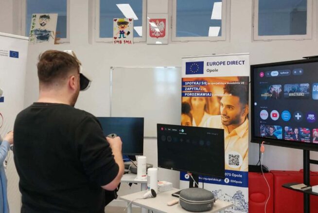 Dwie osoby – kobieta w niebieskim stroju i mężczyzna w czarnej koszulce – mają założone gogle VR i trzymają kontrolery, testując wirtualną rzeczywistość. Stoją przed stanowiskiem z monitorami w sali szkoleniowej. Po prawej stronie widoczny jest duży ekran telewizyjny z interfejsem aplikacji streamingowych i wideo. W tle znajdują się banery promocyjne regionu opolskiego oraz Unii Europejskiej.