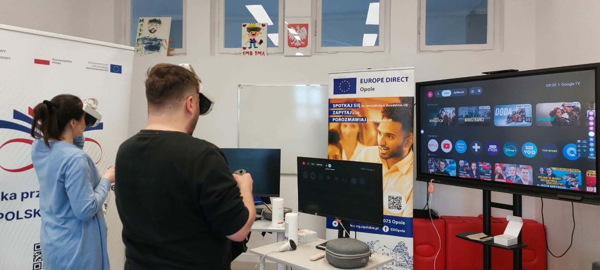 Dwie osoby – kobieta w niebieskim stroju i mężczyzna w czarnej koszulce – mają założone gogle VR i trzymają kontrolery, testując wirtualną rzeczywistość. Stoją przed stanowiskiem z monitorami w sali szkoleniowej. Po prawej stronie widoczny jest duży ekran telewizyjny z interfejsem aplikacji streamingowych i wideo. W tle znajdują się banery promocyjne regionu opolskiego oraz Unii Europejskiej.