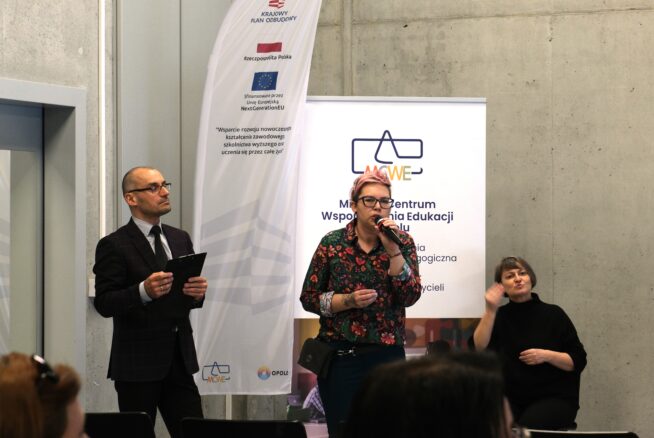 Na zdjęciu troje dorosłych osób prezentuje treści podczas wydarzenia edukacyjnego w nowoczesnej sali. Centralna postać, kobieta w kwiecistej koszuli, przemawia do mikrofonu, a obok niej siedzi inna kobieta tłumacząca wystąpienie na język migowy. Po lewej stronie stoi mężczyzna w garniturze trzymający podkładkę z dokumentami. W tle widoczne są banery informacyjne z logotypami instytucji oraz napisem "Krajowy Plan Odbudowy".