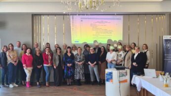 Grupowe zdjęcie wszystkich uczestników konferencji stojących w rzędzie na tle ściany z wyświetloną agendą spotkania. Grupa składa się z kilkudziesięciu osób w różnym wieku, uśmiechających się do aparatu w eleganckim, jasnym wnętrzu z dużym żyrandolem.