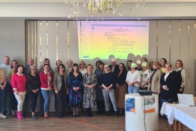 Grupowe zdjęcie wszystkich uczestników konferencji stojących w rzędzie na tle ściany z wyświetloną agendą spotkania. Grupa składa się z kilkudziesięciu osób w różnym wieku, uśmiechających się do aparatu w eleganckim, jasnym wnętrzu z dużym żyrandolem.