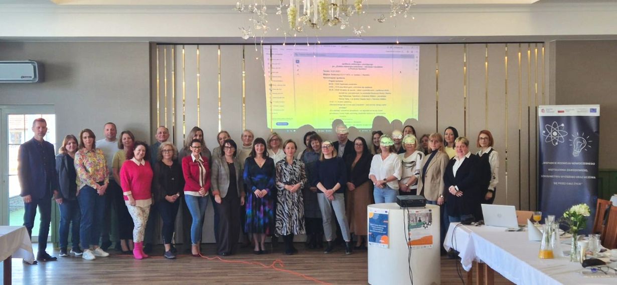 Grupowe zdjęcie wszystkich uczestników konferencji stojących w rzędzie na tle ściany z wyświetloną agendą spotkania. Grupa składa się z kilkudziesięciu osób w różnym wieku, uśmiechających się do aparatu w eleganckim, jasnym wnętrzu z dużym żyrandolem.