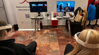Dwóch uczestników siedzi w goglach VR przed stanowiskiem z dwoma monitorami, na których widać symulacje różnych sytuacji życiowych. Między nimi znajduje się szklany stolik z akcesoriami technicznymi oraz baner promocyjny województwa opolskiego. W tle widać fragmenty garderoby i materiały szkoleniowe.