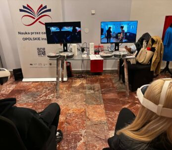 Dwóch uczestników siedzi w goglach VR przed stanowiskiem z dwoma monitorami, na których widać symulacje różnych sytuacji życiowych. Między nimi znajduje się szklany stolik z akcesoriami technicznymi oraz baner promocyjny województwa opolskiego. W tle widać fragmenty garderoby i materiały szkoleniowe.