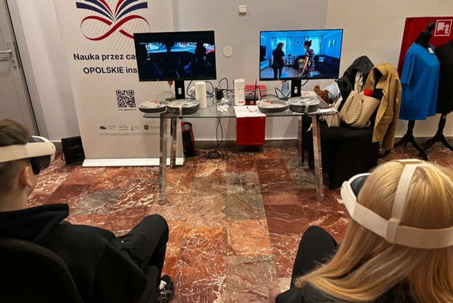 Dwóch uczestników siedzi w goglach VR przed stanowiskiem z dwoma monitorami, na których widać symulacje różnych sytuacji życiowych. Między nimi znajduje się szklany stolik z akcesoriami technicznymi oraz baner promocyjny województwa opolskiego. W tle widać fragmenty garderoby i materiały szkoleniowe.