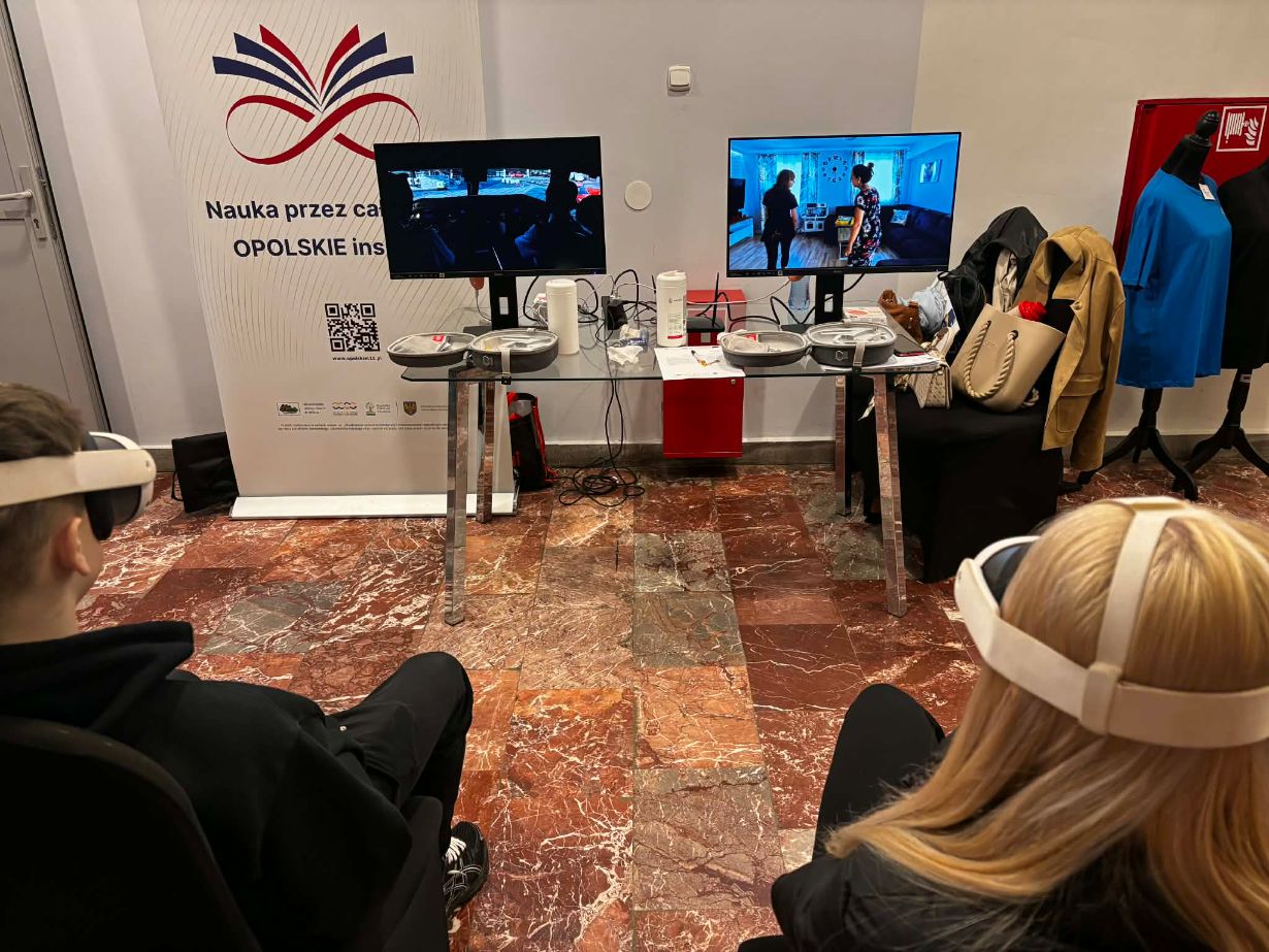 Dwóch uczestników siedzi w goglach VR przed stanowiskiem z dwoma monitorami, na których widać symulacje różnych sytuacji życiowych. Między nimi znajduje się szklany stolik z akcesoriami technicznymi oraz baner promocyjny województwa opolskiego. W tle widać fragmenty garderoby i materiały szkoleniowe.