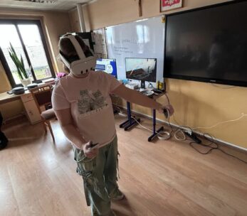 Dziewczyna w różowej koszulce z kotami i szerokich spodniach testuje zestaw VR w centrum klasy. Trzyma kontrolery i wykonuje gesty rękami, zanurzona w wirtualnej rzeczywistości. W tle widać monitory, duży ekran telewizyjny oraz okno, przez które wpada naturalne światło. Na tablicy zapisany jest adres strony internetowej projektu.