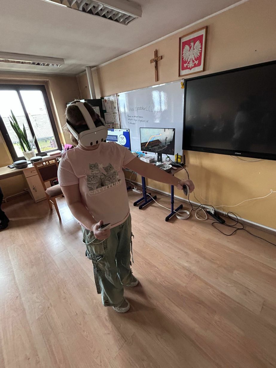Dziewczyna w różowej koszulce z kotami i szerokich spodniach testuje zestaw VR w centrum klasy. Trzyma kontrolery i wykonuje gesty rękami, zanurzona w wirtualnej rzeczywistości. W tle widać monitory, duży ekran telewizyjny oraz okno, przez które wpada naturalne światło. Na tablicy zapisany jest adres strony internetowej projektu.