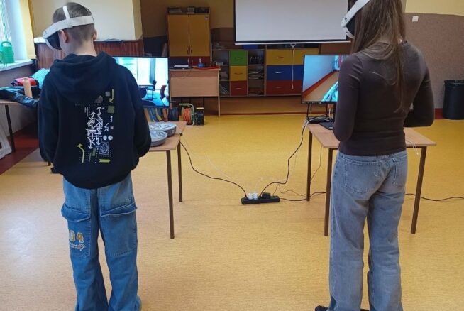 Szerokie ujęcie sali, w której dwie osoby stoją tyłem, korzystając z zestawów do rzeczywistości wirtualnej. Każda z nich ma przed sobą osobne stanowisko z monitorem podłączonym do gogli. W tle znajduje się biały ekran projekcyjny, kolorowe szafki oraz krzyż zawieszony nad drzwiami.
