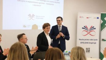 Mężczyzna w granatowym garniturze i kobieta w czarnym żakiecie stoją na podium przed ekranem projekcyjnym. Mężczyzna trzyma mikrofon, a kobieta patrzy w stronę publiczności, która bije brawo. Po prawej stronie widoczny jest biały roll-up z kolorową grafiką i hasłem 