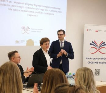 Mężczyzna w granatowym garniturze i kobieta w czarnym żakiecie stoją na podium przed ekranem projekcyjnym. Mężczyzna trzyma mikrofon, a kobieta patrzy w stronę publiczności, która bije brawo. Po prawej stronie widoczny jest biały roll-up z kolorową grafiką i hasłem "Nauka przez całe życie – OPOLSKIE inspiruje".
