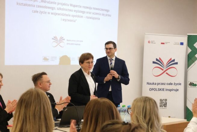Mężczyzna w granatowym garniturze i kobieta w czarnym żakiecie stoją na podium przed ekranem projekcyjnym. Mężczyzna trzyma mikrofon, a kobieta patrzy w stronę publiczności, która bije brawo. Po prawej stronie widoczny jest biały roll-up z kolorową grafiką i hasłem "Nauka przez całe życie – OPOLSKIE inspiruje".