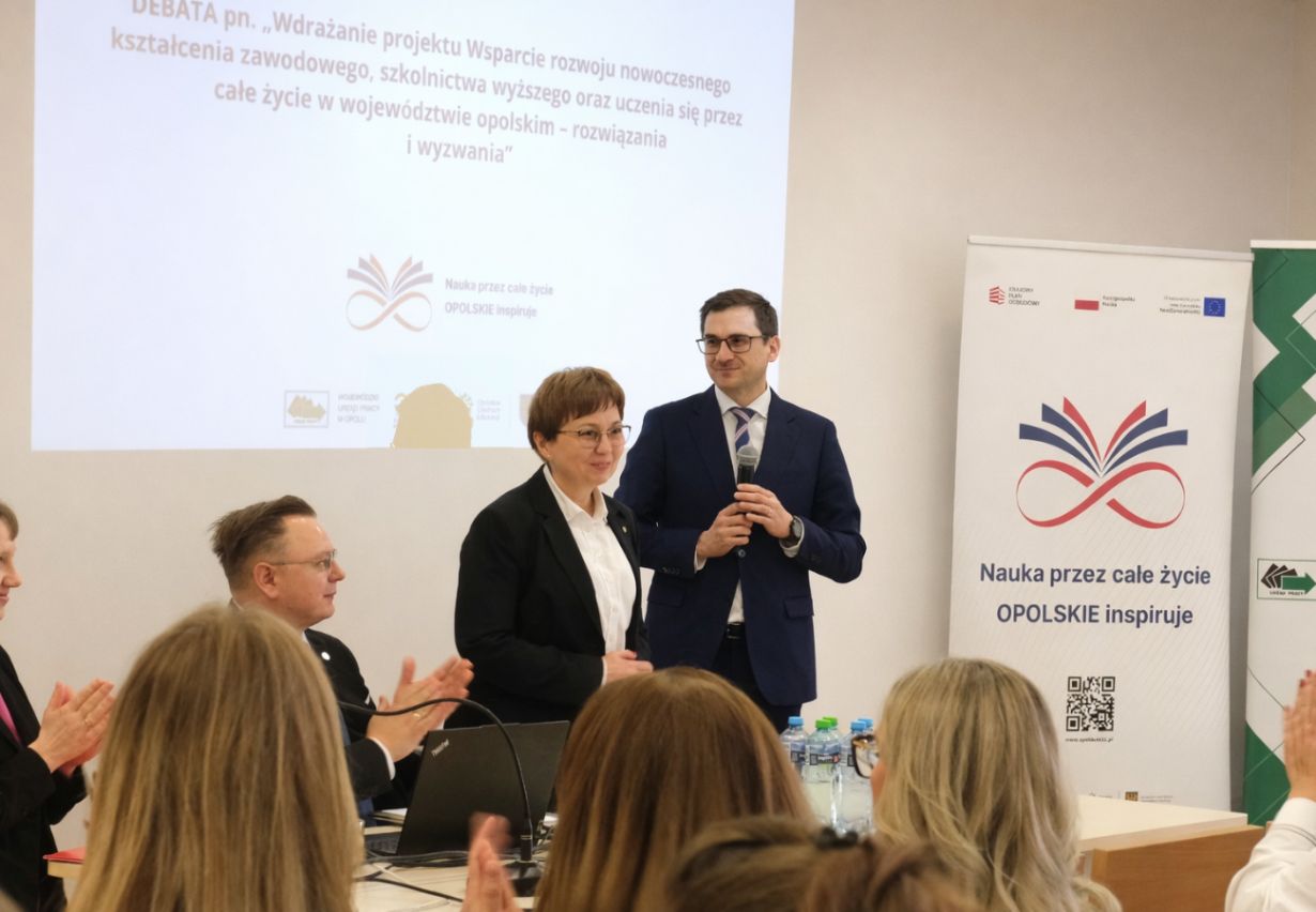 Mężczyzna w granatowym garniturze i kobieta w czarnym żakiecie stoją na podium przed ekranem projekcyjnym. Mężczyzna trzyma mikrofon, a kobieta patrzy w stronę publiczności, która bije brawo. Po prawej stronie widoczny jest biały roll-up z kolorową grafiką i hasłem 