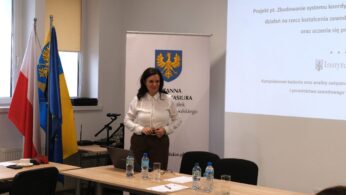 Portretowe ujęcie Wicemarszałek Zuzanny Donath-Kasiury stojącej przy ekranie z prezentacją projektu „Instytutu Śląskiego”. Prelegentka ubrana jest w białą koszulę i brązowe spodnie, a jej postawa sugeruje aktywny dialog z uczestnikami. Na stole przed nią stoją butelki z wodą mineralną i laptop.